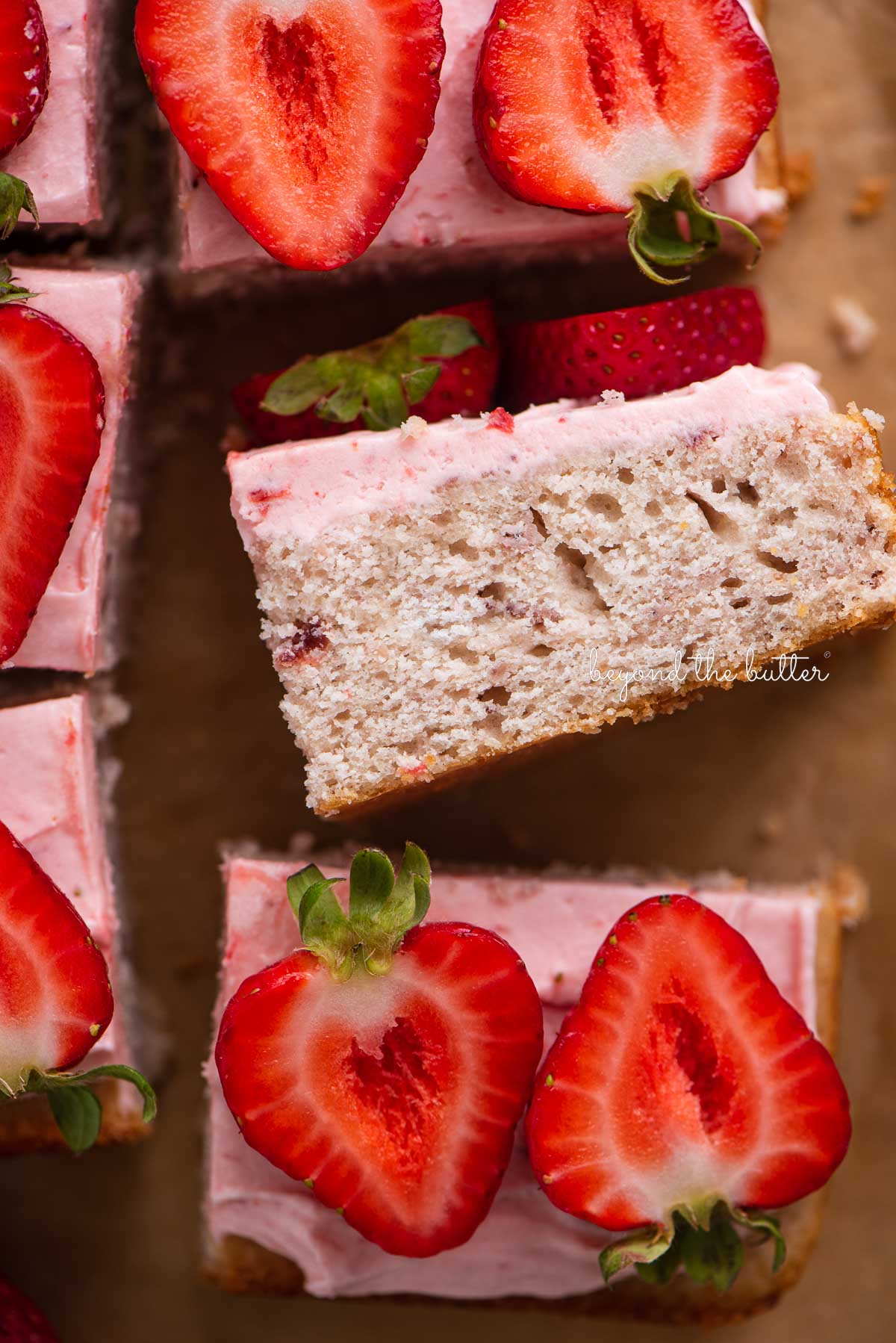 Slices of mini strawberry sheet cake topped with strawberry buttercream frosting and fresh strawberries with one slice on its side | © Beyond the Butter®