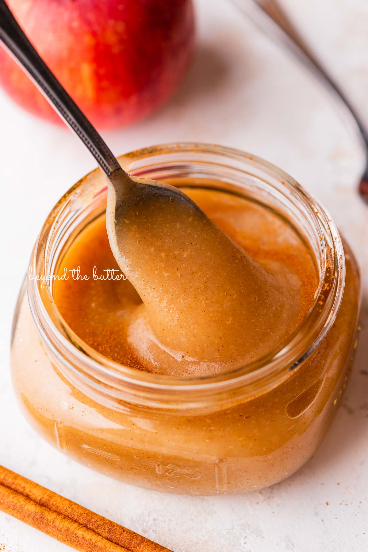 Filled glass jar of no sugar added homemade applesauce with a spoon on white background with apples.