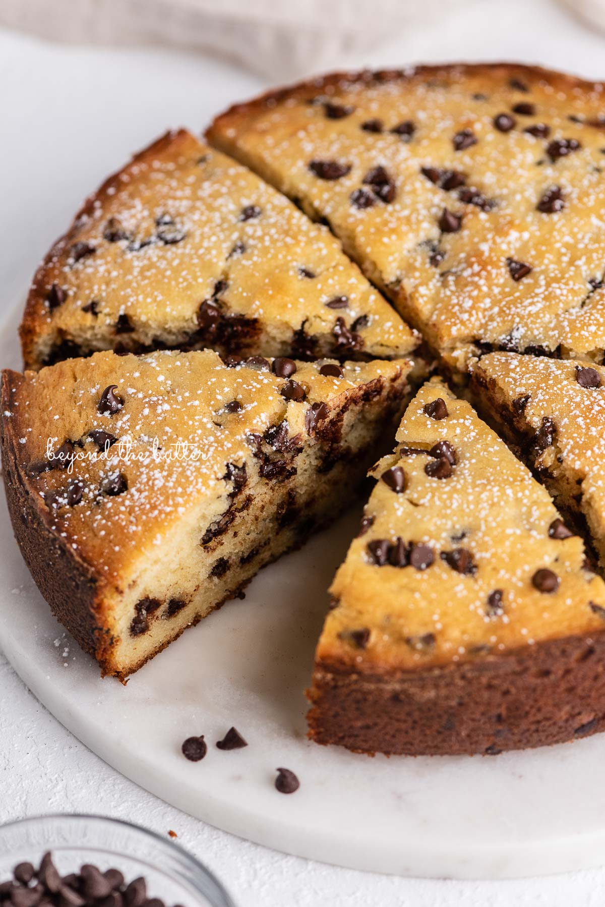 Sliced chocolate chip ricotta cake on a marble cake round with small bowl of mini chocolate chips nearby | © Beyond the Butter®