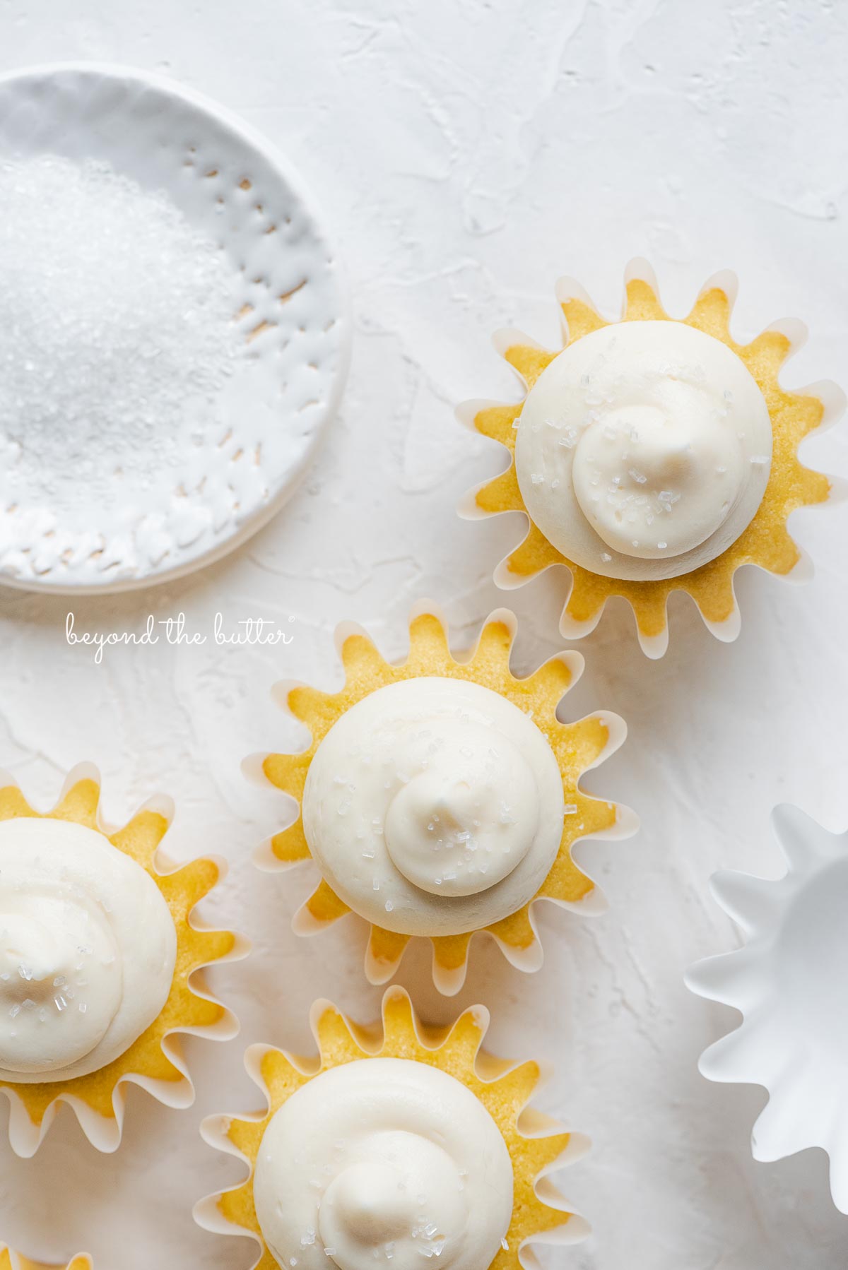 Overhead image of frosted vanilla cupcakes sprinkled with sparkling sugar randomly placed on white background next to empty cupcake liner and small shallow bowl of white sparkling sugar.
