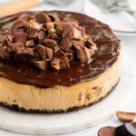 Peanut butter cheesecake topped with chocolate ganache and mini Reese's peanut butter cups on marble plate on white background.