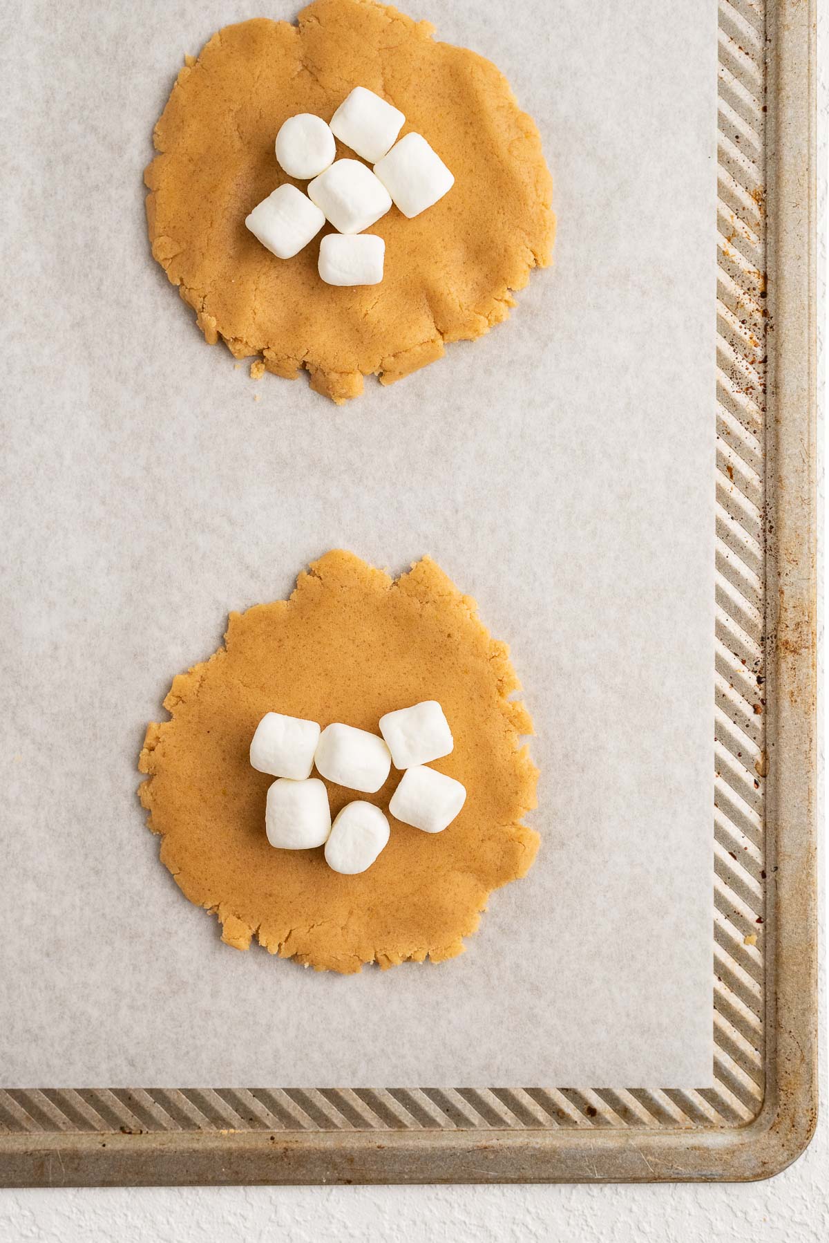 Six mini marshmallows placed on fluffernutter cookie discs on parchment paper lined baking sheet.