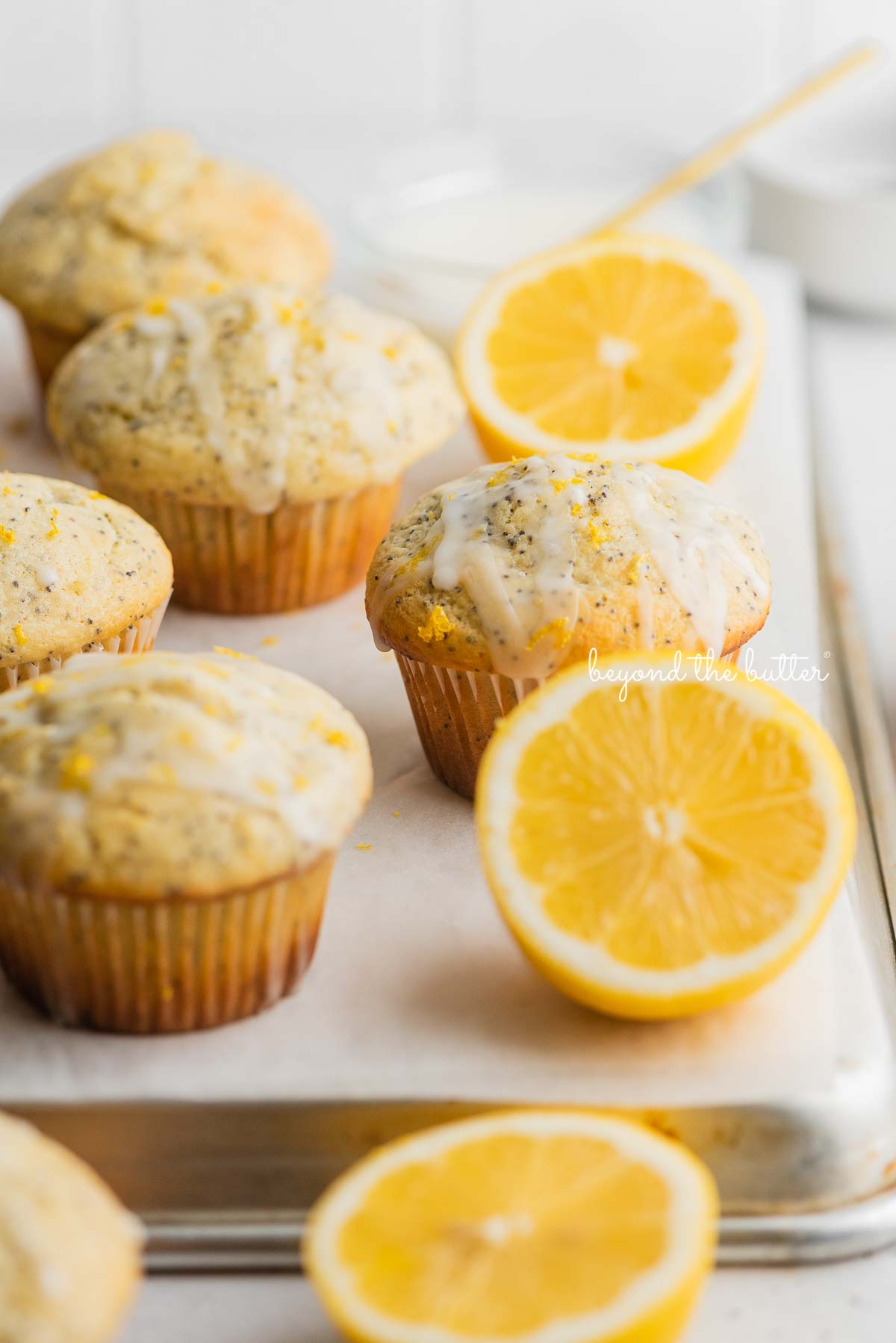 Lemon poppyseed muffins randomly placed on a white parchment paper inverted baking sheet with bowl of glaze and half slices of lemons next to them.