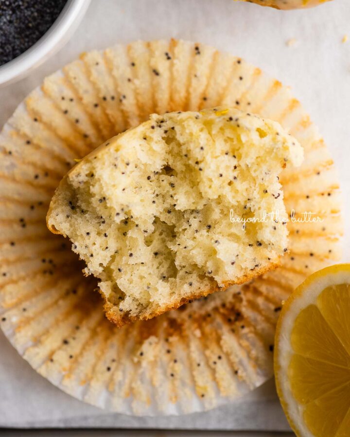 Half-eaten lemon poppy seed muffin on top of muffin liner on white background with a bowl of poppy seeds and slice of lemon.