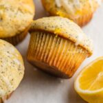 Randomly placed lemon poppy seed muffins on a parchment paper-lined baking sheet with half of a lemon.