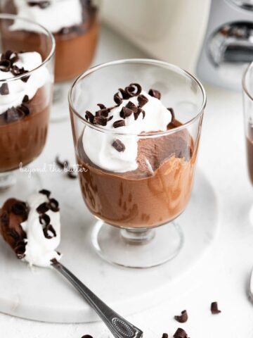 Parfait goblets filled with chocolate mousse, whipped cream, and chocolate curls with a spoonful laying next to one on a white marble slab and white textured background.