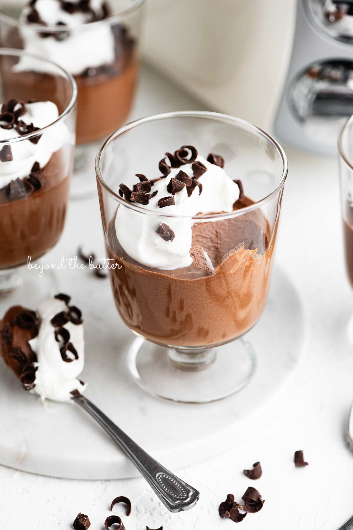 Parfait goblets filled with chocolate mousse, whipped cream, and chocolate curls with a spoonful laying next to one on a white marble slab and white textured background.