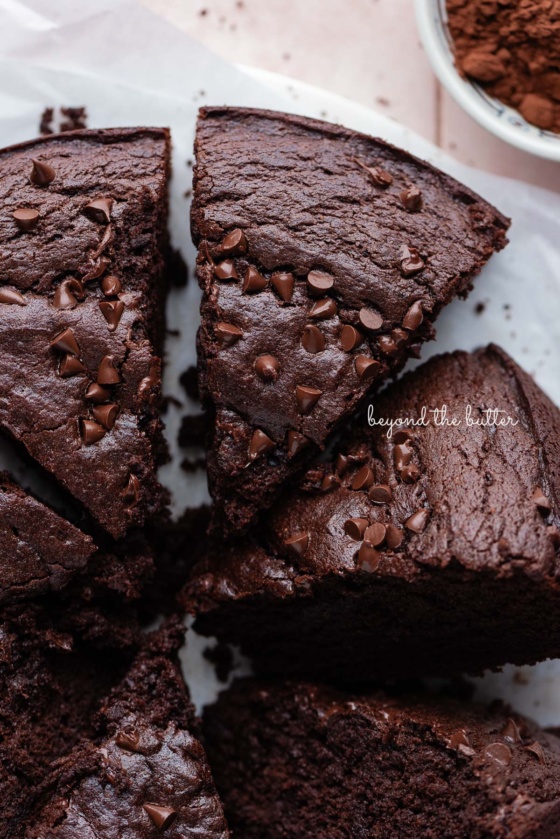 Slices of chocolate ricotta cake topped with chocolate chips on white parchment paper and a small bowl of cocoa powder placed on a light pink tiled background.
