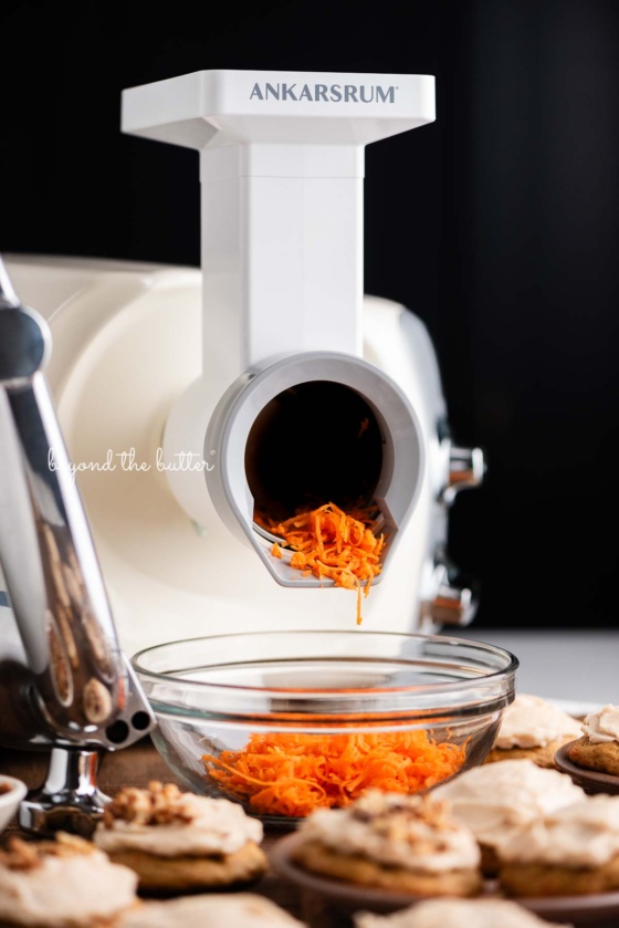 Light creme color ankarsrum mixer using the vegetable cutter attachment to grate carrots into a glass bowl for carrot cake cookies against a dark background.