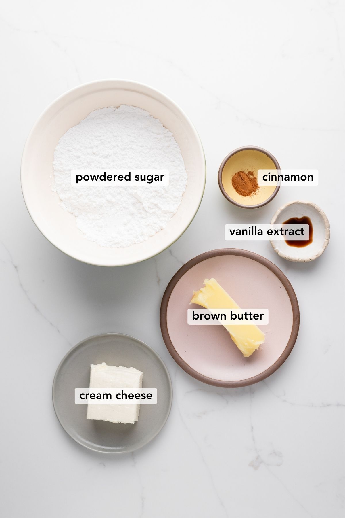 Brown butter cream cheese frosting ingredients in various size bowls labeled as powdered sugar, cinnamon, vanilla extract, brown butter, and cream cheese on a white marbled background.