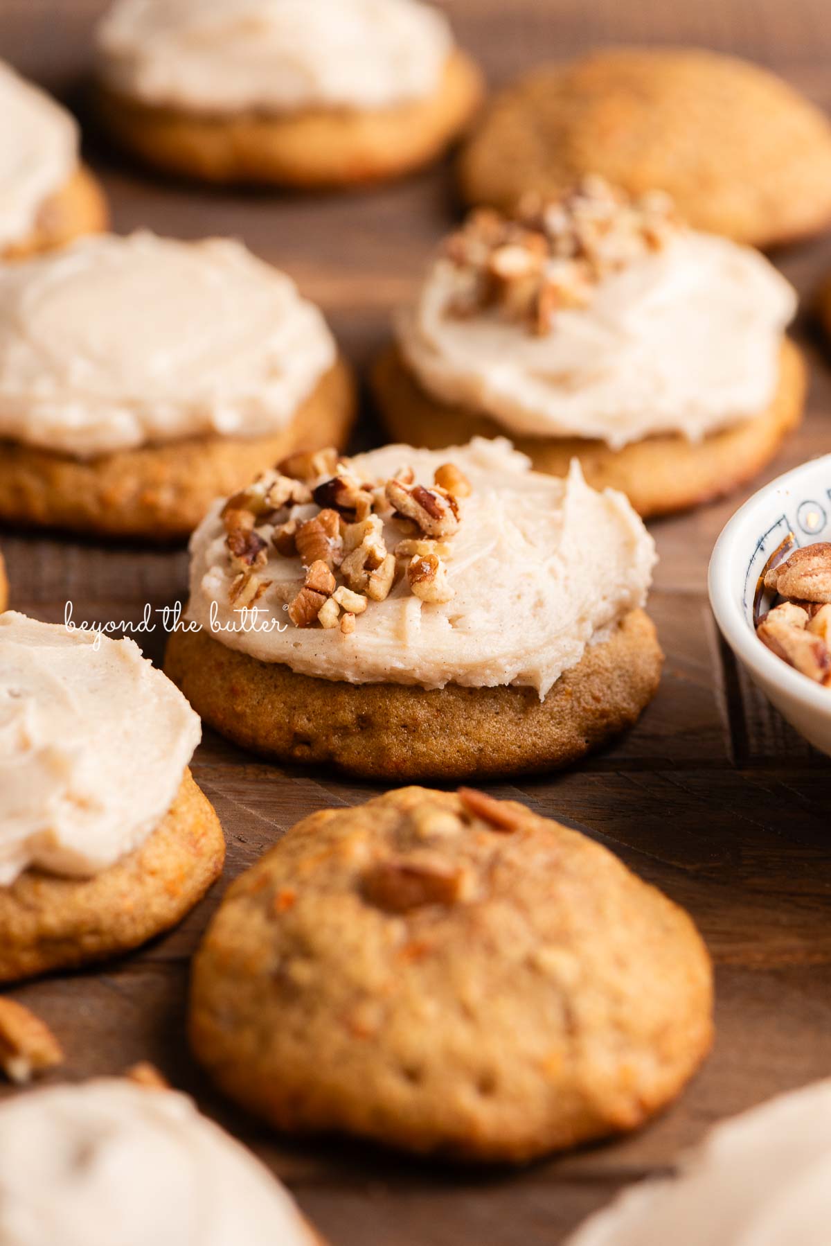 Carrot cake cookies frosted with brown butter cream cheese frosting and some topped with chopped pecans with a small bowl filled with chopped pecans to the side on a dark wood background.