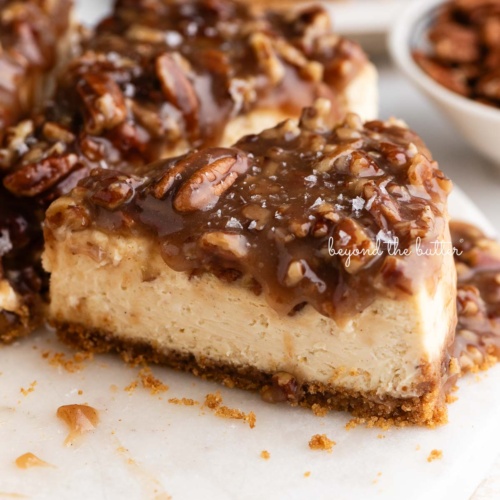 Sliced pecan pie cheesecake sprinkled with sea salt flakes on a white marble cake stand placed next to a small bowl of pecans on a white background.