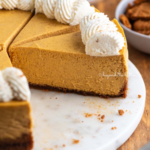 Sliced pumpkin cheesecake topped with piped whipped cream on a white marbled cake plate on a dark wood background.