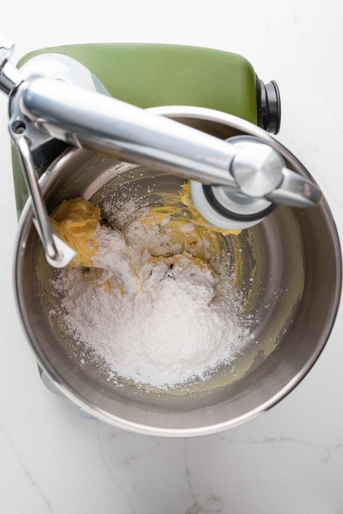 Powdered sugar and beatened unsalted butter in a forest green Ankarsrum Assistent Original Mixer on a white marbled surface.