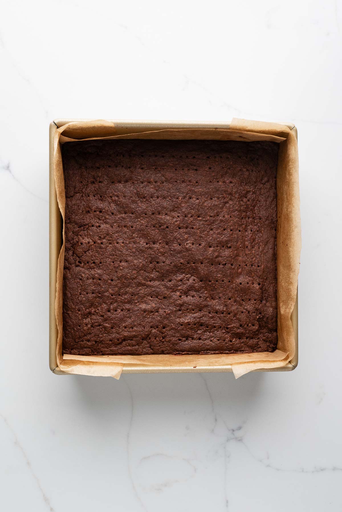 Baked chocolate shortbread biscuits in a parchment paper lined metal baking tin on a white marbled surface.