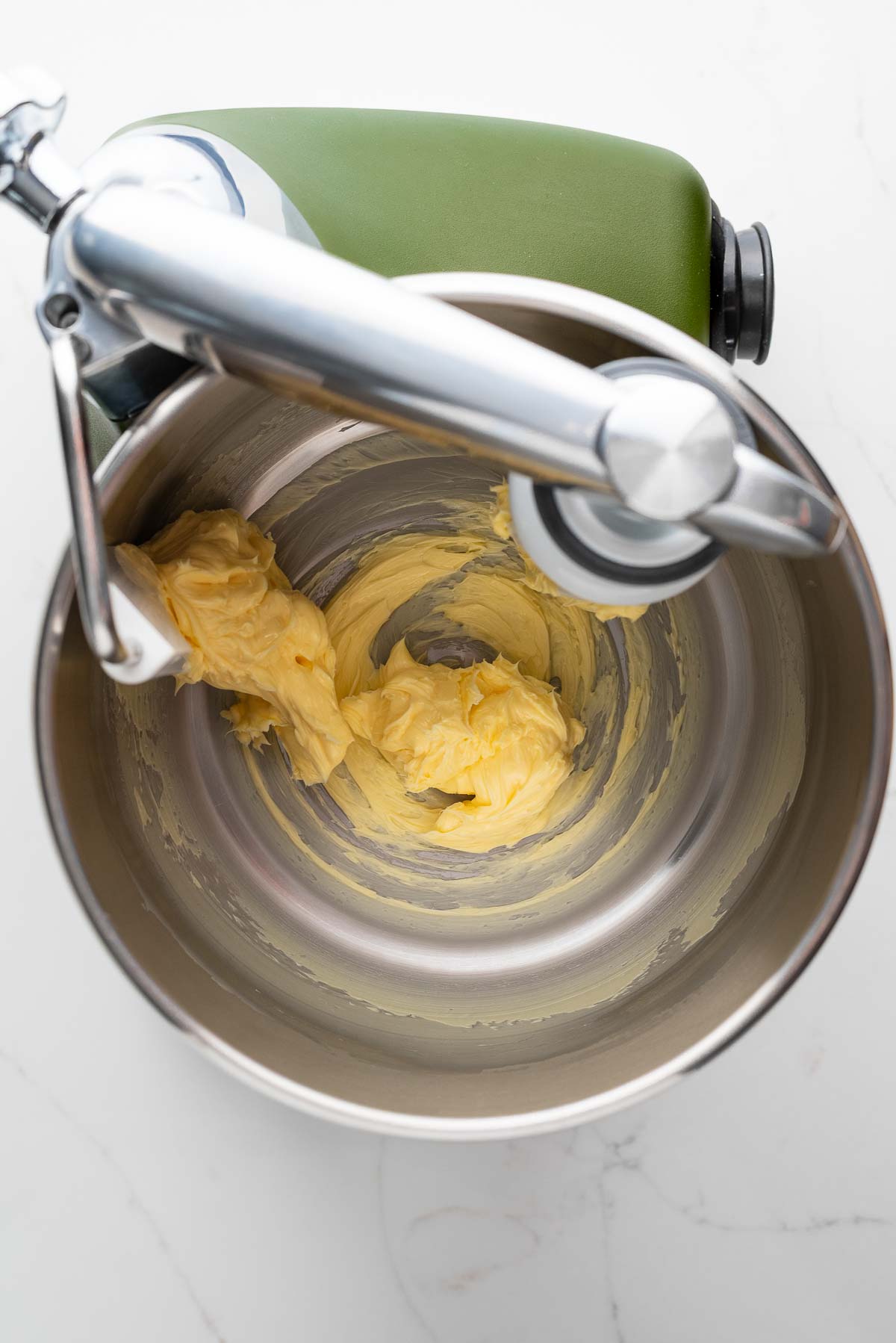 Beating unsalted butter in a forest green colored Ankarsrum Assistent Original Mixer on a white marbled surface.