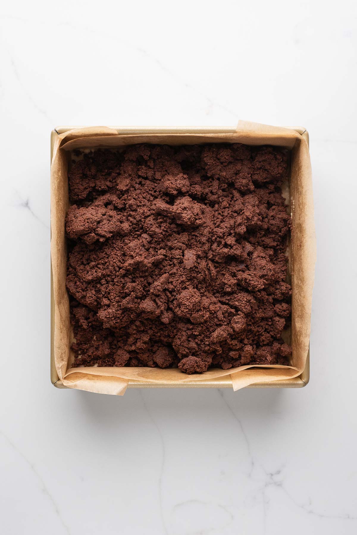 Chocolate shortbread dough transferred to a parchment paper lined eight inch square baking tin on a white marbled surface.