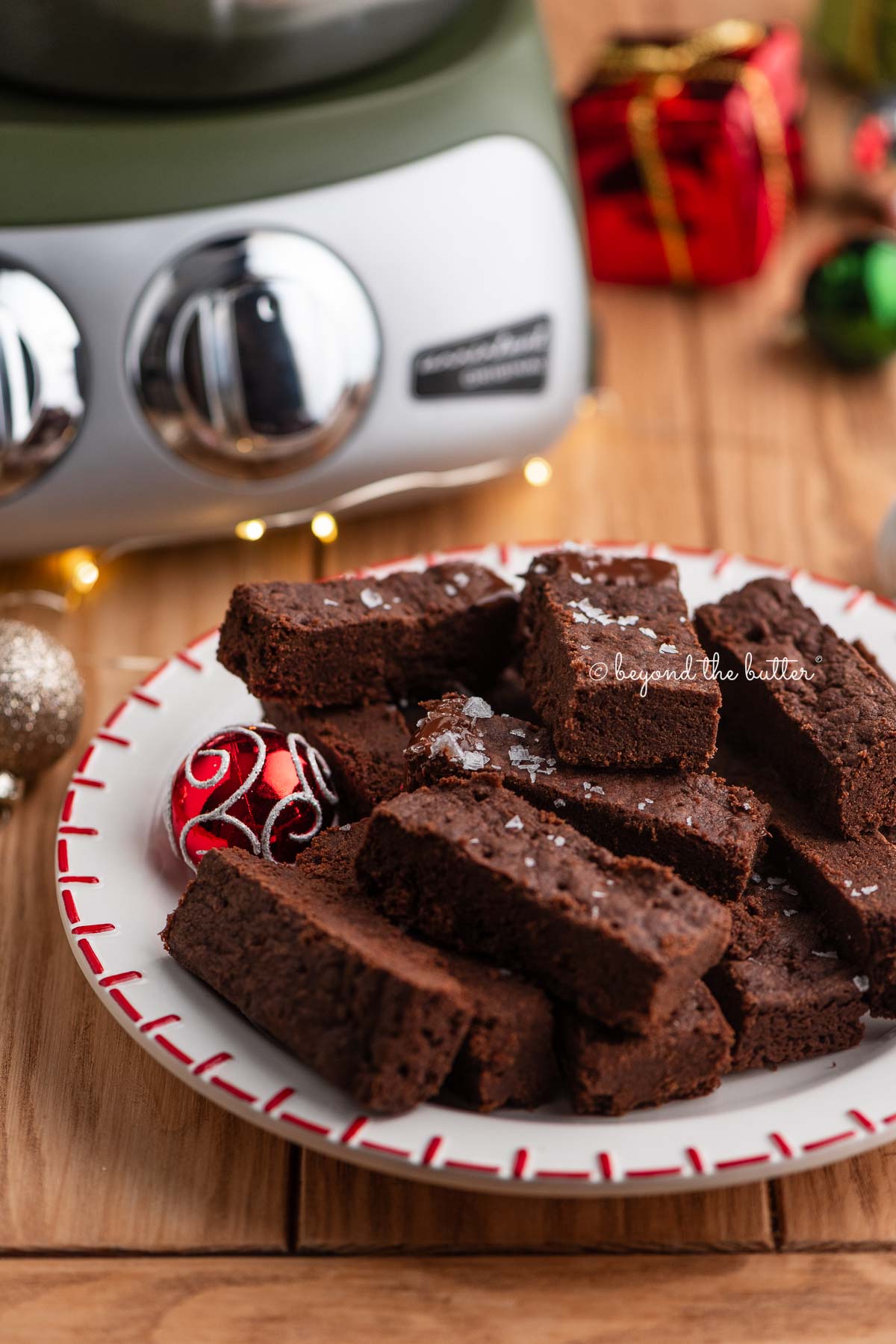 Red and white dessert plate topped with chocolate shortbread surrounded by miniature ball ornaments, fairy lights, and a forest green Ankarsrum Assistent Original mixer on a natural wood background.