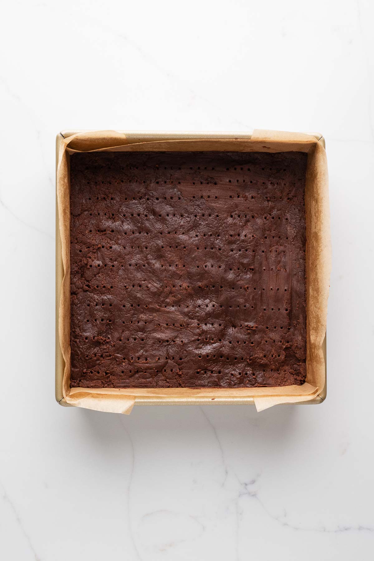 Chocolate shortbread dough pressed into an eight inch parchment paper lined baking tin docked on top using a fork.