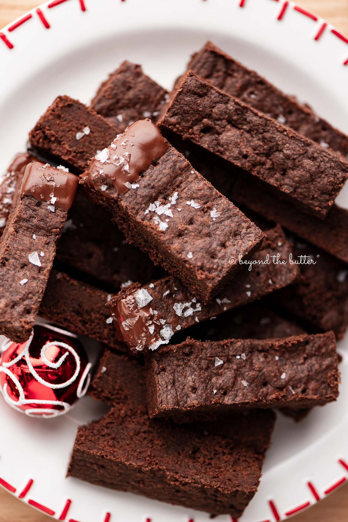 Sliced chocolate shortbread biscuits sprinkled with flaky sea salt flakes and placed on a round red and white holiday-themed dessert plate surrounded by miniature ball ornaments on a wood surface.