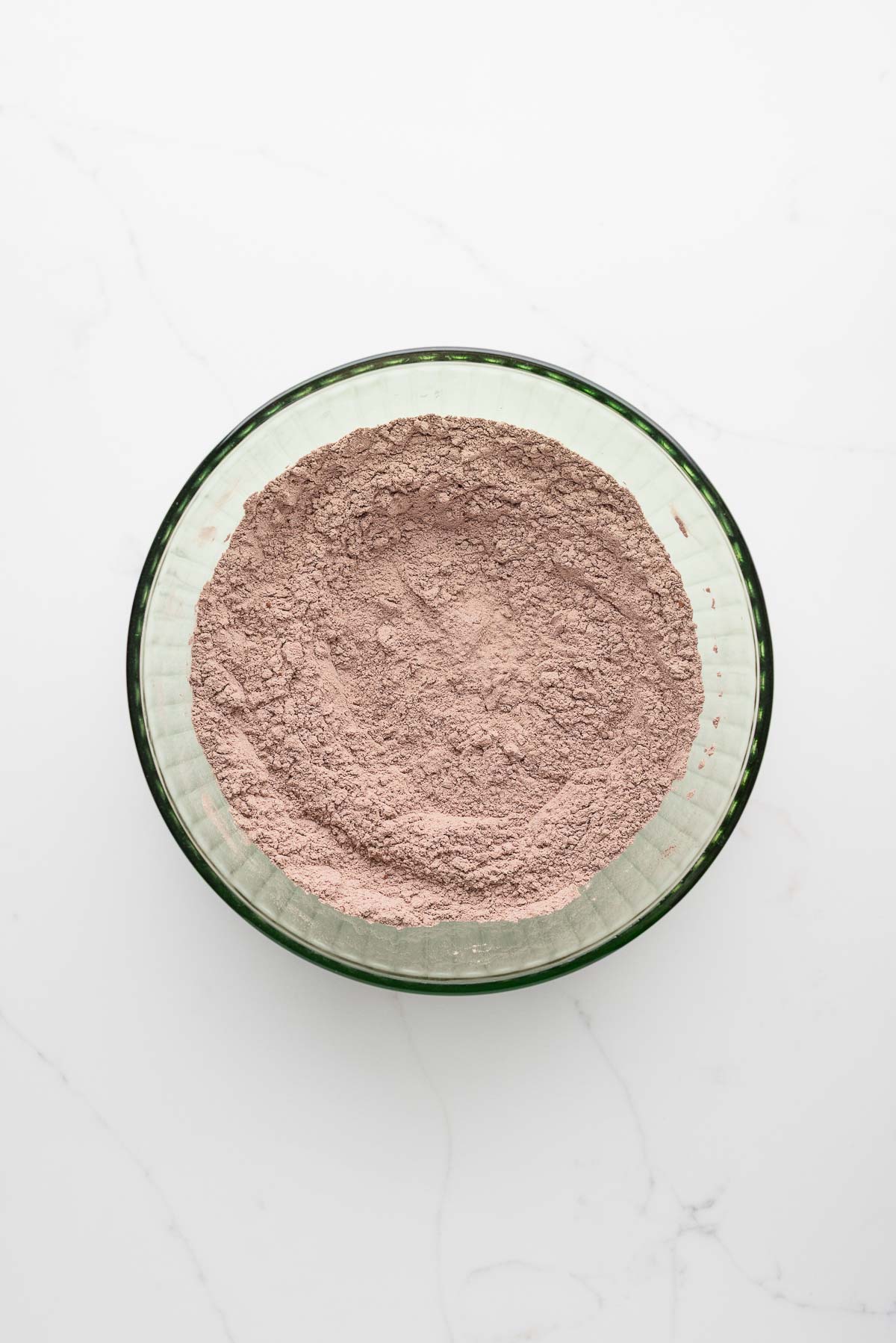 Whisked flour, dutch process cocoa powder, baking soda, and salt in a green glass mixing bowl on a white marbled surface.