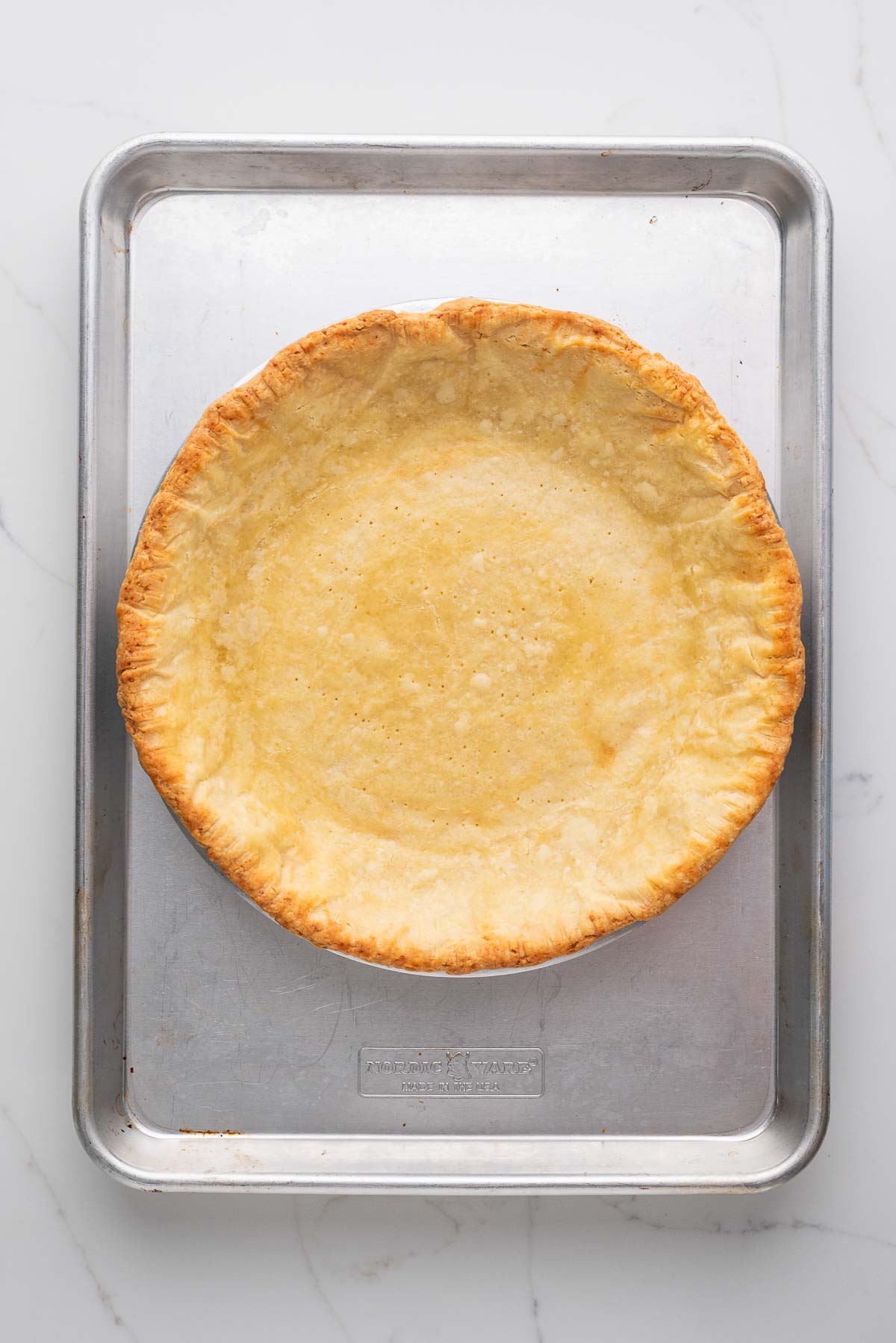Blind baked pie crust in a metal pie in placed on a metal baking sheet and white marbled background.