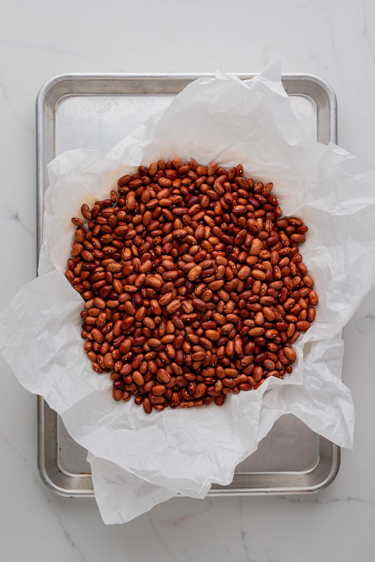 Homemade pie crust placed on a metal baking sheet topped with overlapping sheets of crumpled parchment paper and dried beans for pie weights.