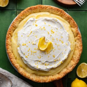 Lemon cream pie topped with fresh whipped cream and lemon zest centered on a green tiled background surrounded by lemons, wood dessert plates, forks, napkin, and pie server.