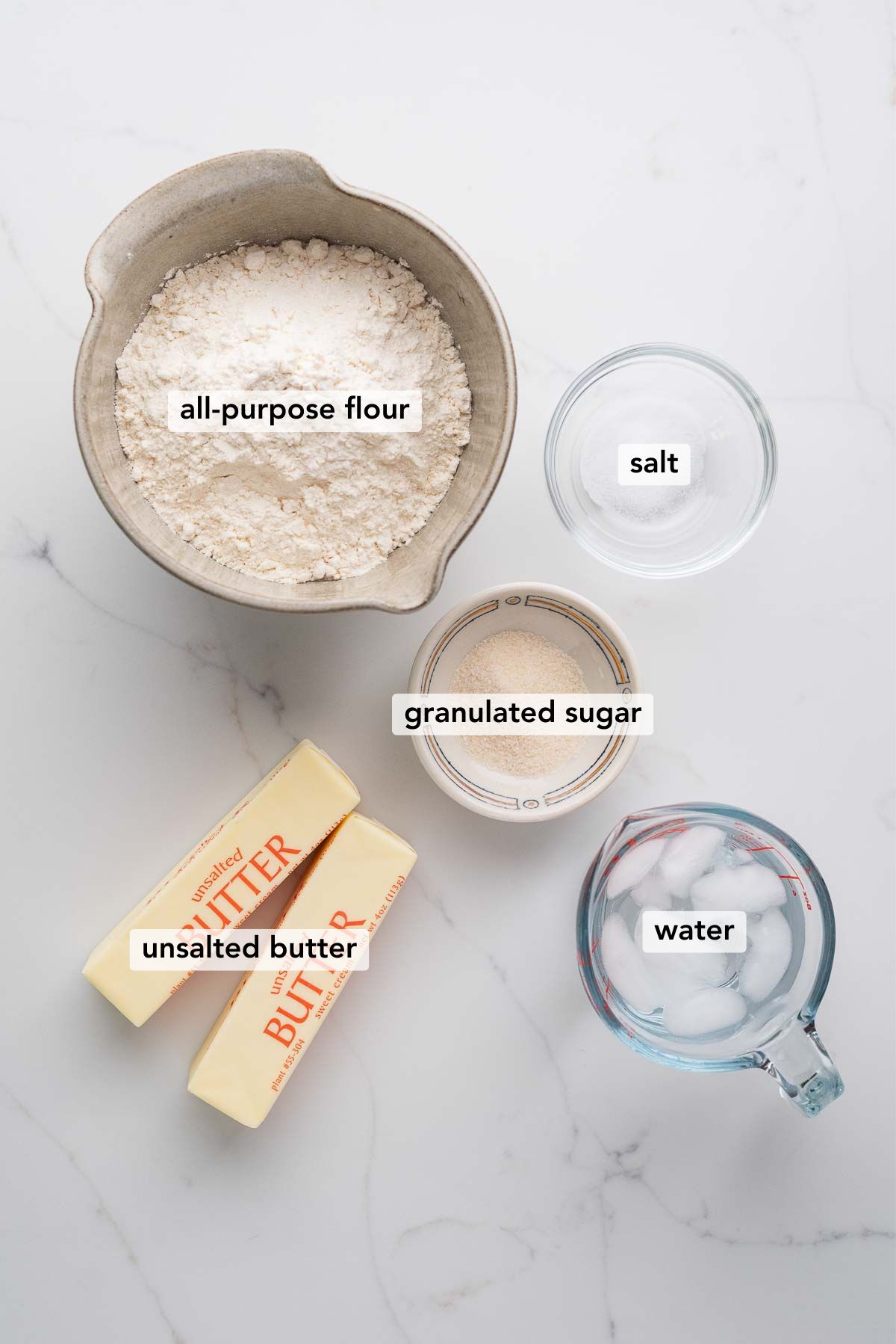 Ingredients in various size bowls labeled as all purpose flour, salt, granulated sugar, unsalted butter, and water on a white marbled background.
