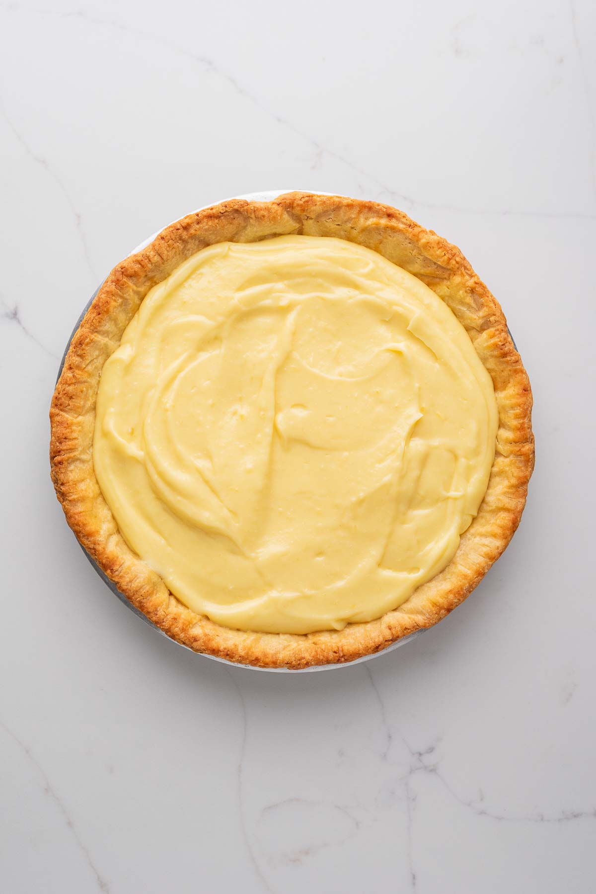 Lemon cream pie filling in a blind baked pie crust on a white marbled background.