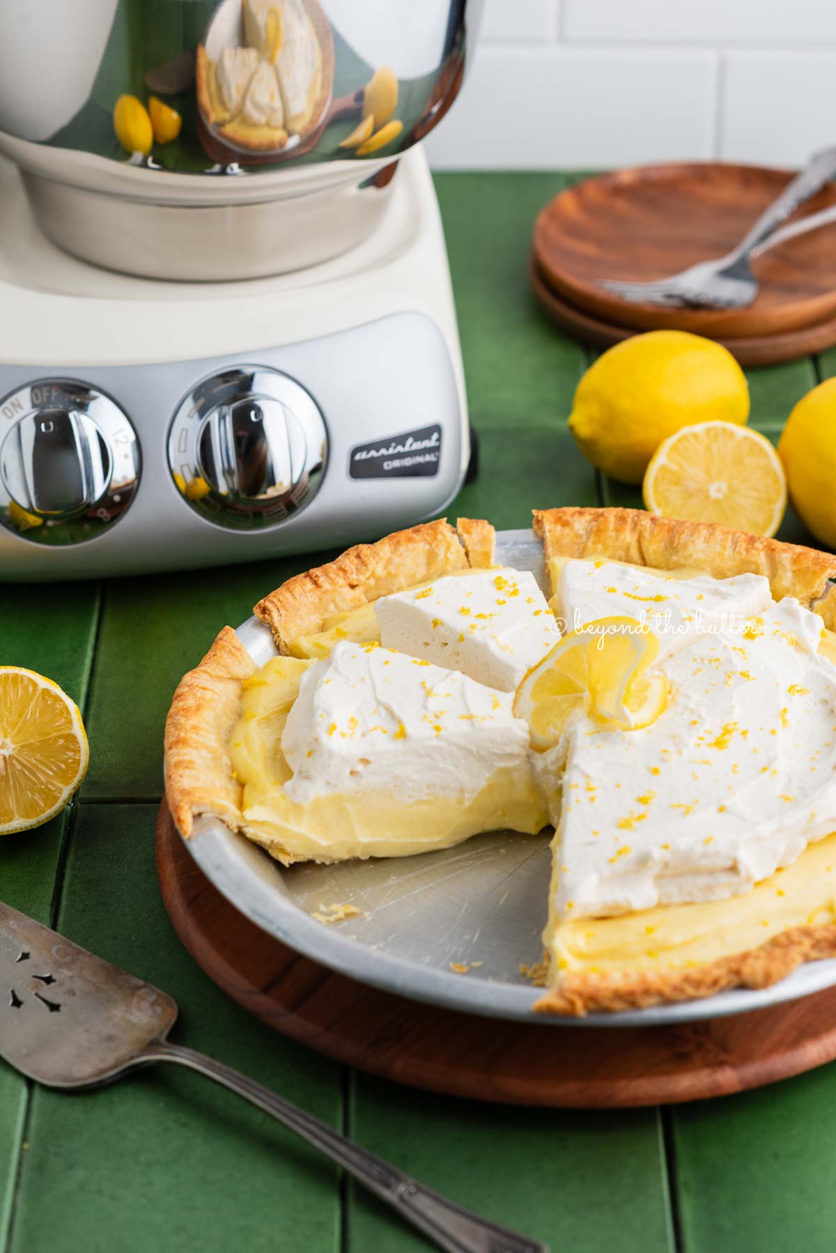 Sliced lemon sour cream pie on a wooden pie serving board next to a creme light colored ankarsrum assistent original mixer, lemons, pie server, and dessert plates.