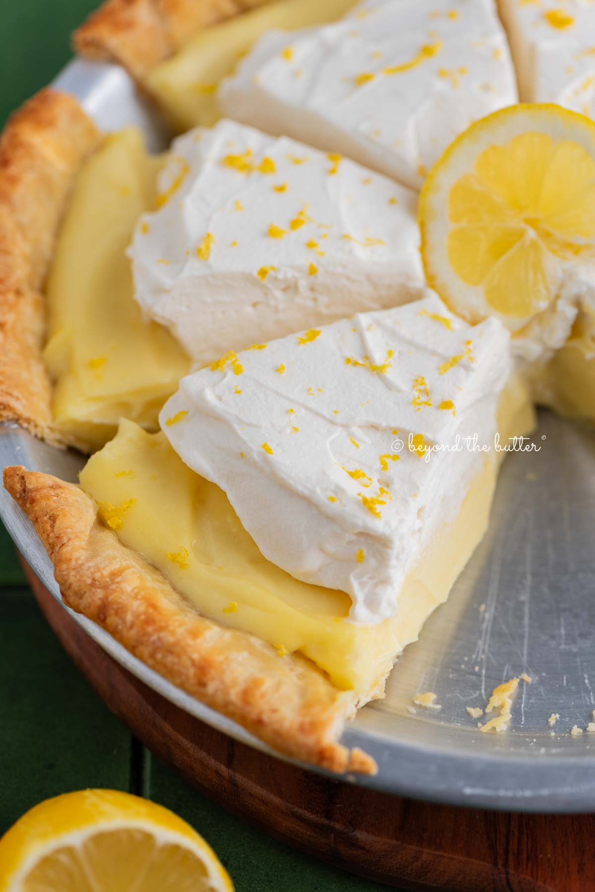 Slices of lemon sour cream pie topped with whipped cream and lemon zest in a metal pie tin on a green tiled background.