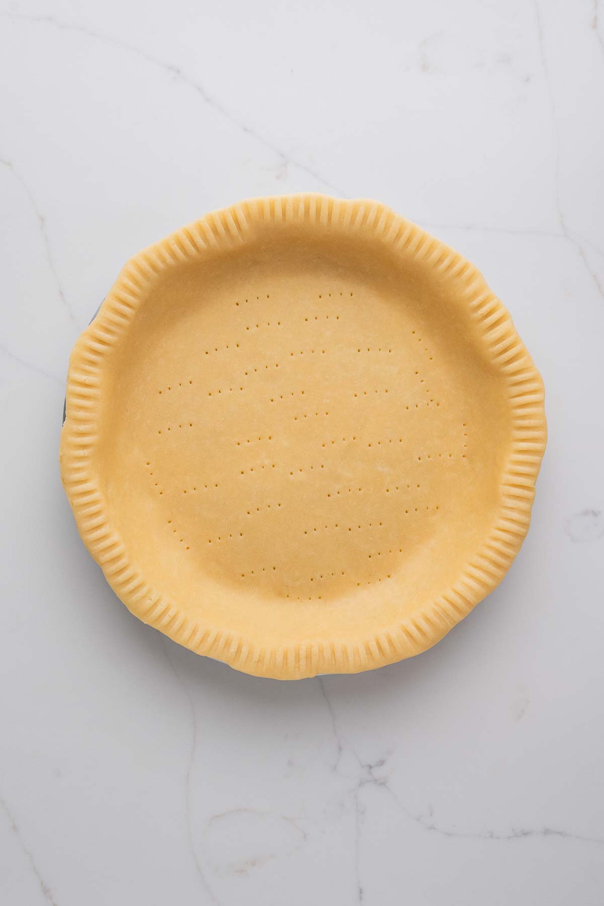 Rolled out pie dough placed in a metal pie tin that's been trimmed, edges crimped and bottom docked with a fork on a white marbled background.