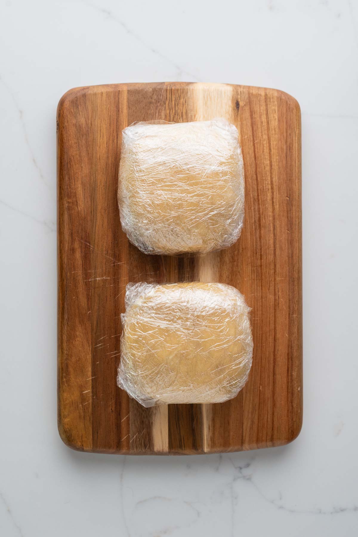 Two pie dough discs double wrapped in plastic wrap placed on a wood cutting board on a white marbled background.