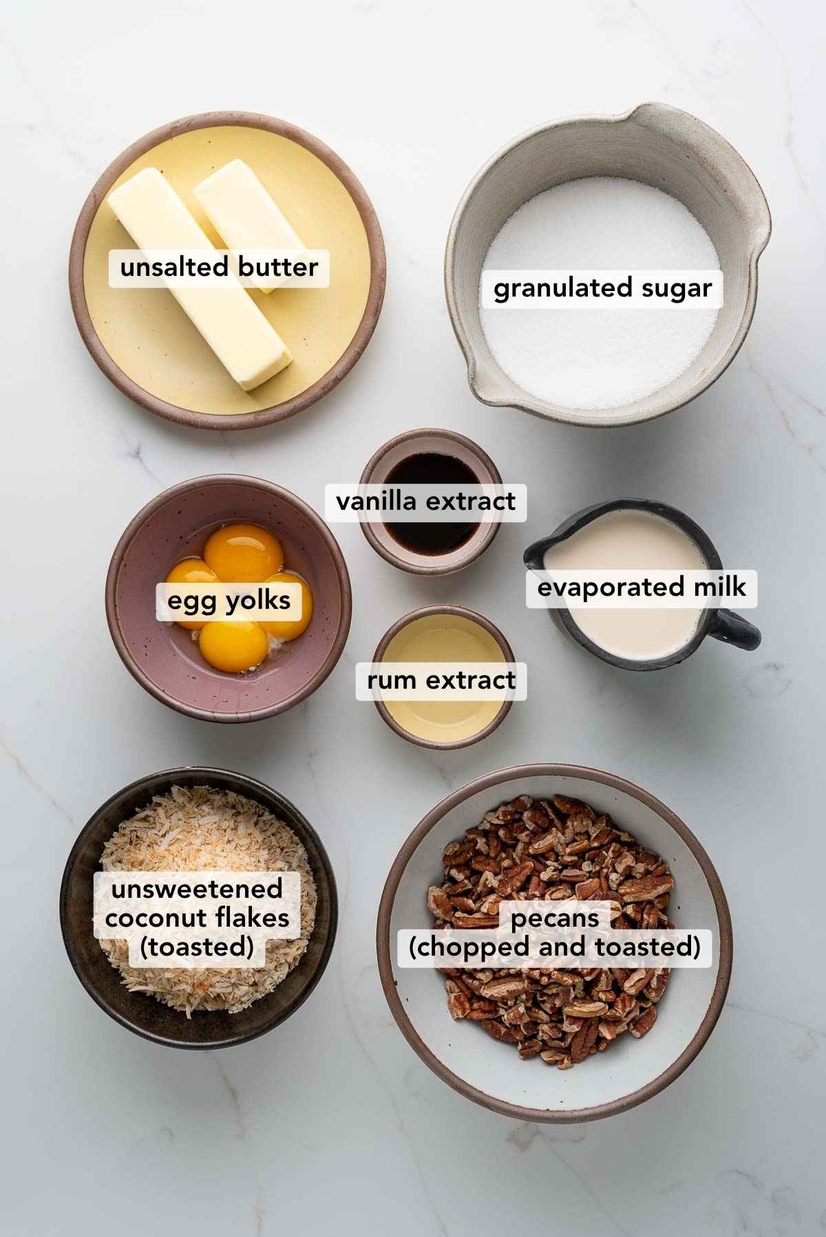 Ingredients in various size bowls labeled as unsalted butter, granulated sugar, egg yolks, vanilla extract, rum extract, evaporated milk, unsweetened coconut flakes (toasted), and pecans (chopped and toasted) on a white marbled background.
