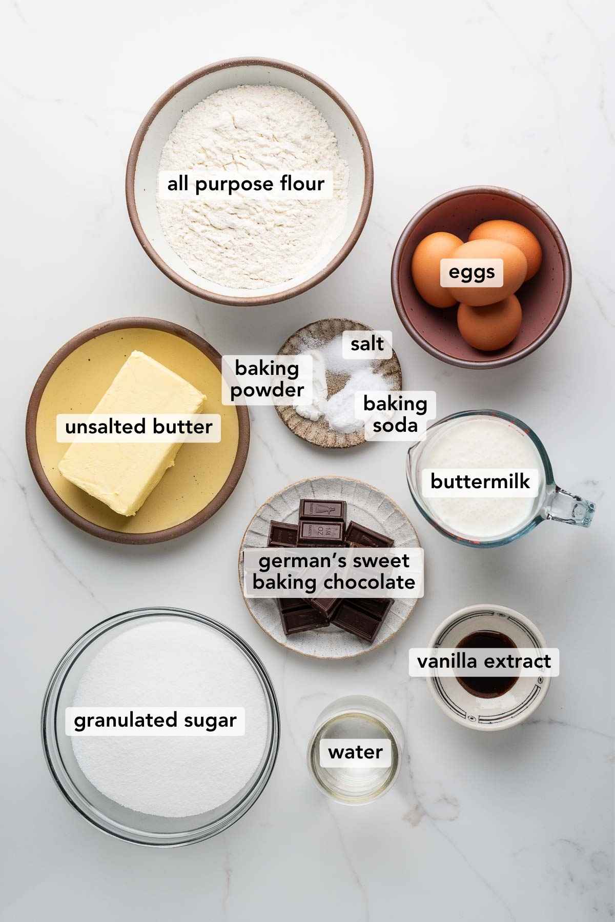 Ingredients in various size bowls labeled as all purpose flour, eggs, unsalted butter, baking powder, baking soda, salt, buttermilk, german's sweet baking chocolate, vanilla extract, water, and granulated sugar on a white marbled background.