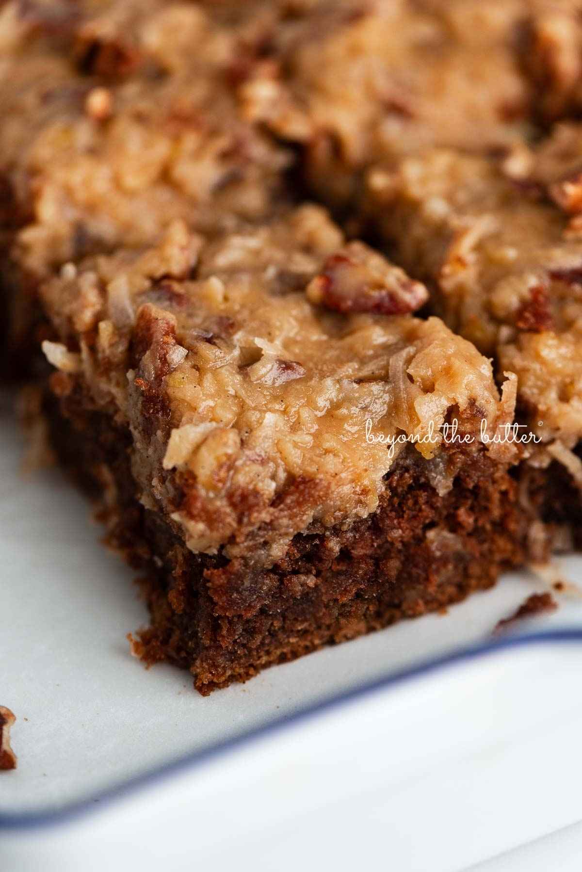 Sliced German chocolate cake with coconut pecan frosting in a parchment paper lined ceramic baking pan.