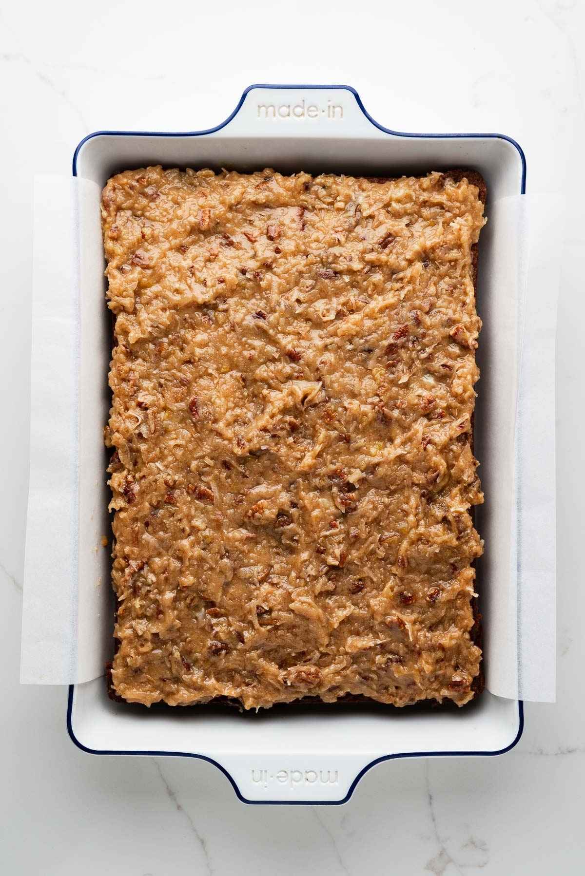 Coconut pecan frosted German chocolate sheet cake in a parchment paper lined white ceramic baking dish on a white marbled background.