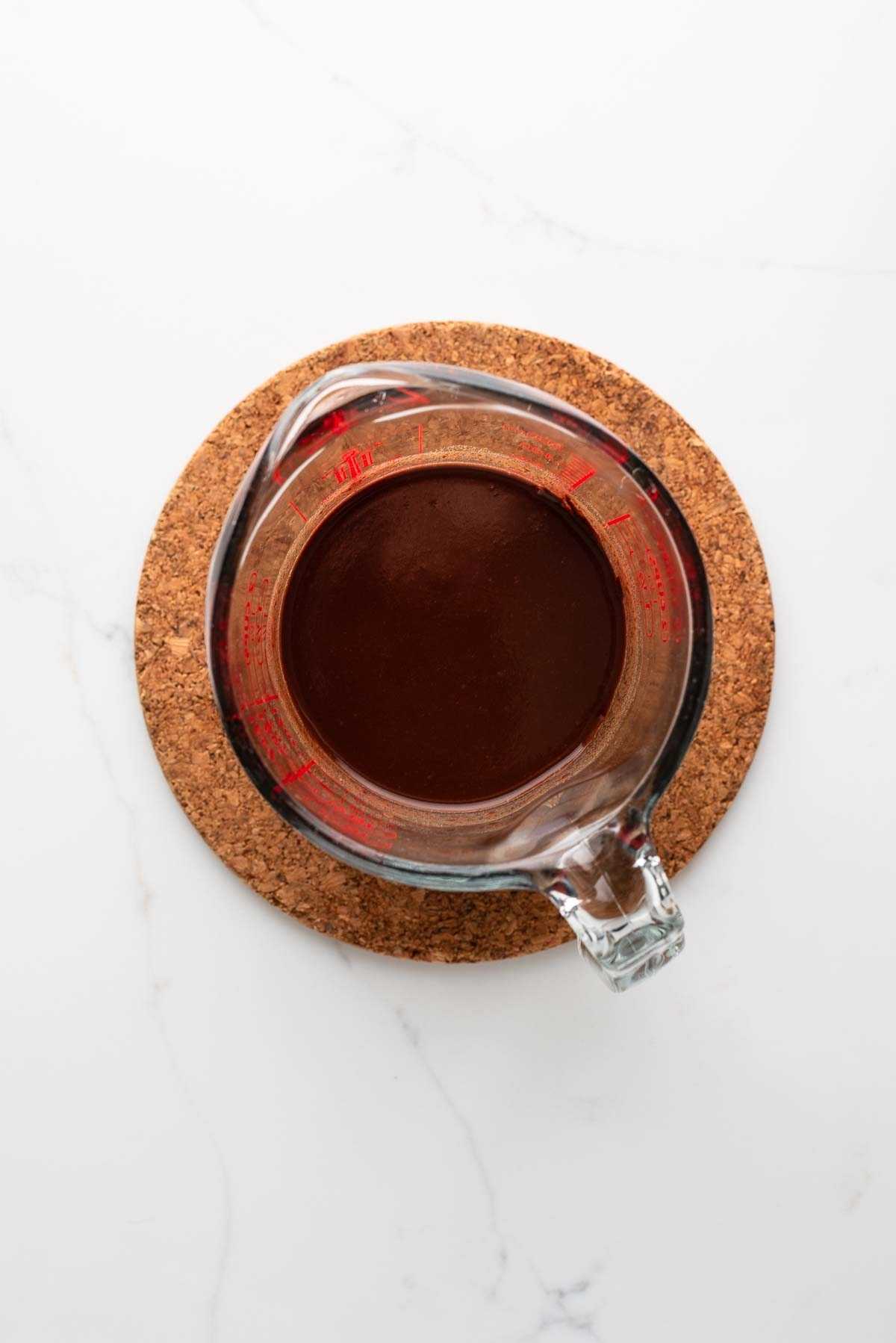 Melted chocolate and water together in a glass measuring cup sitting on a cork trivet on a white marbled background.