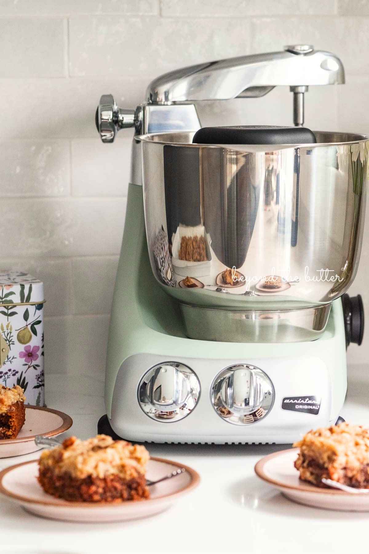 Ankarsrum assistent original mixer in nordic sage on white quartz kitchen counter with subway tile backsplash with recipe box and dessert plates with german chocolate sheet cake around it.