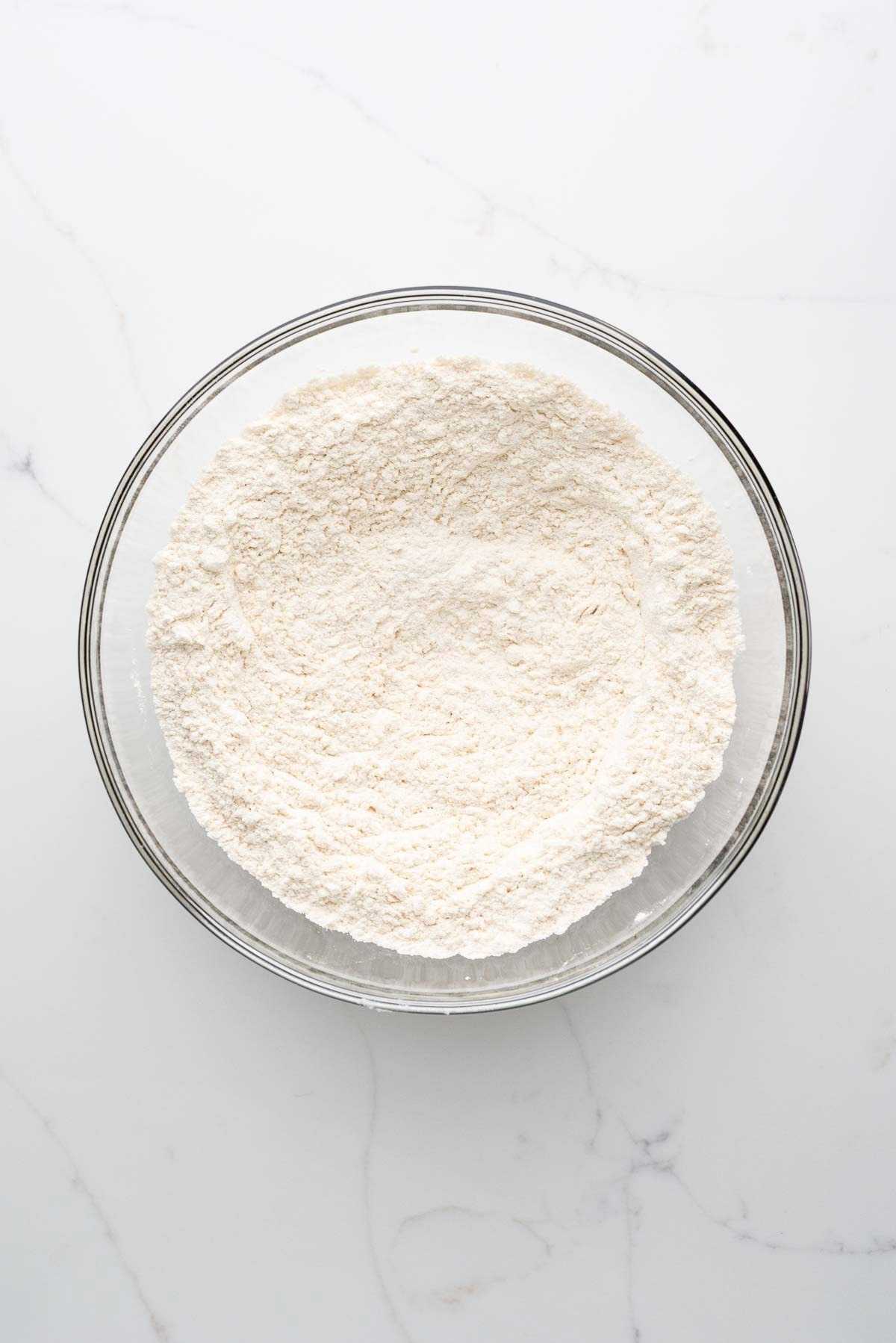 Whisked all purpose flour, baking soda, baking powder, and salt in a medium-sized glass bowl on a white marbled background.