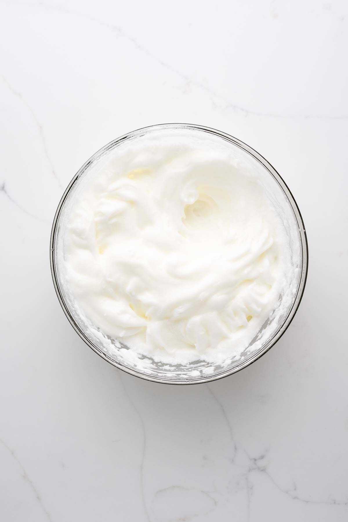 Egg whites whisked to stiff peaks in a small glass bowl on a white marbled background.